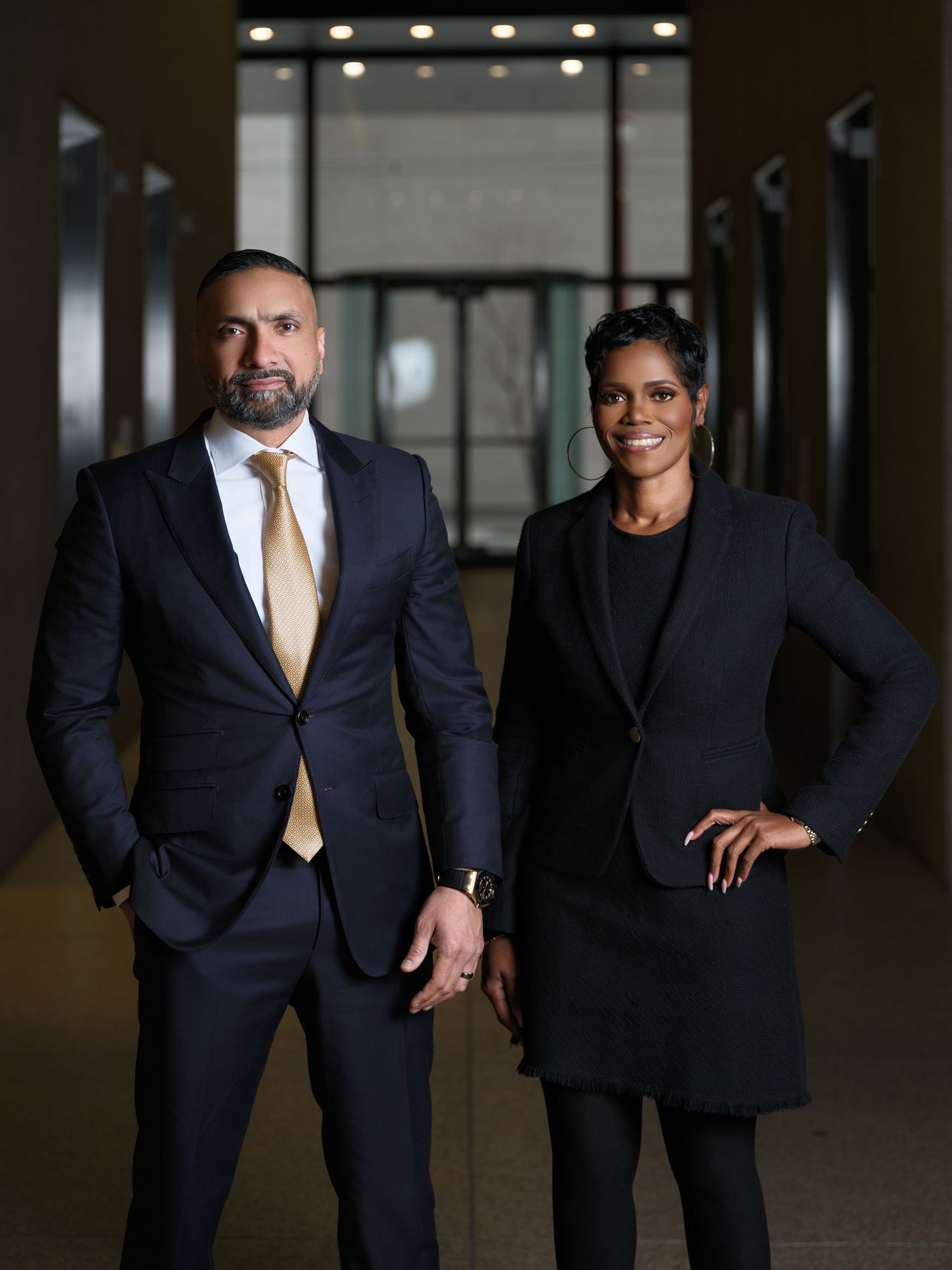 Two business professionals in dark suits pose confidently in modern office hallway.