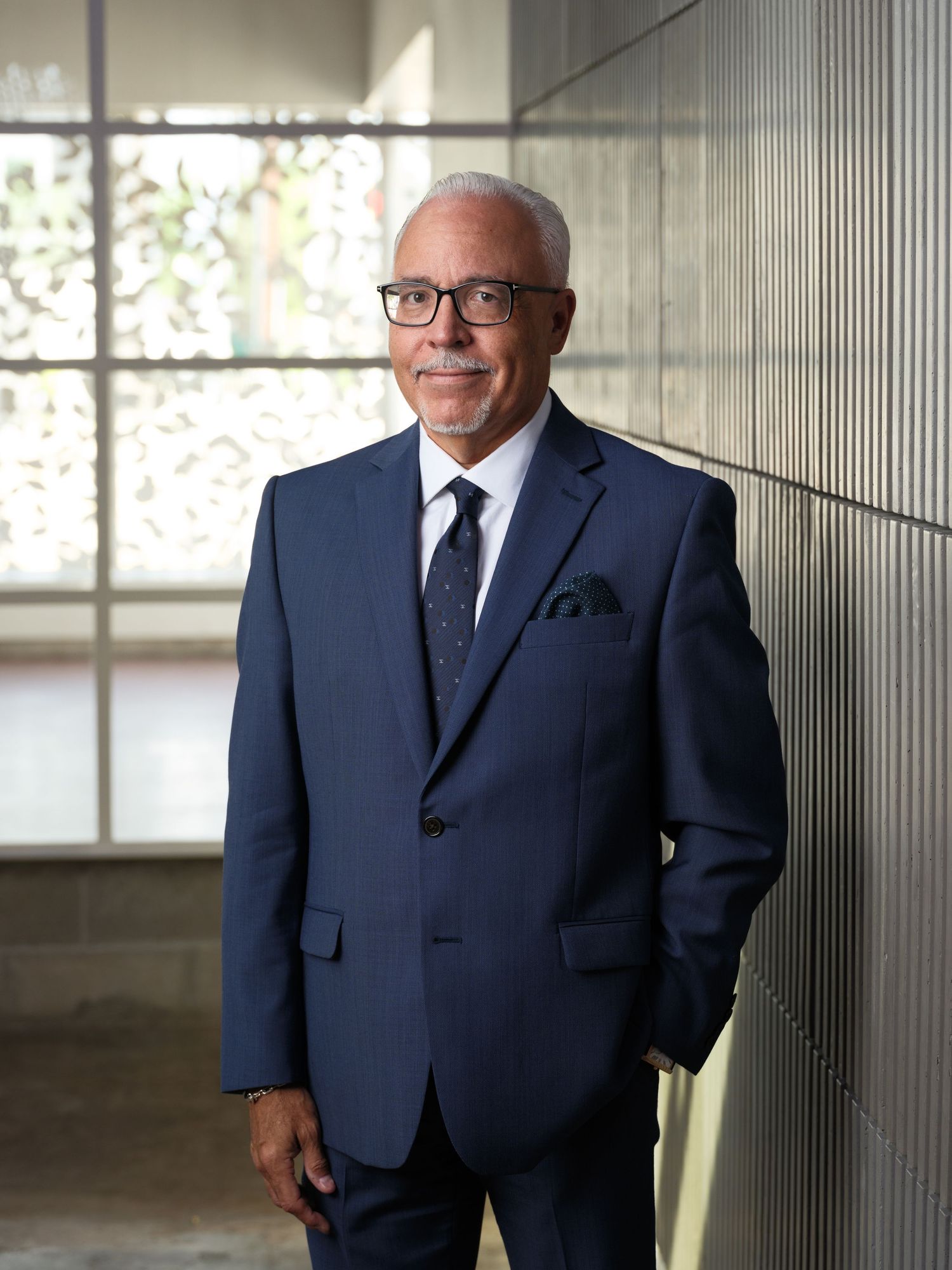 Distinguished executive in navy suit standing by window with natural lighting.