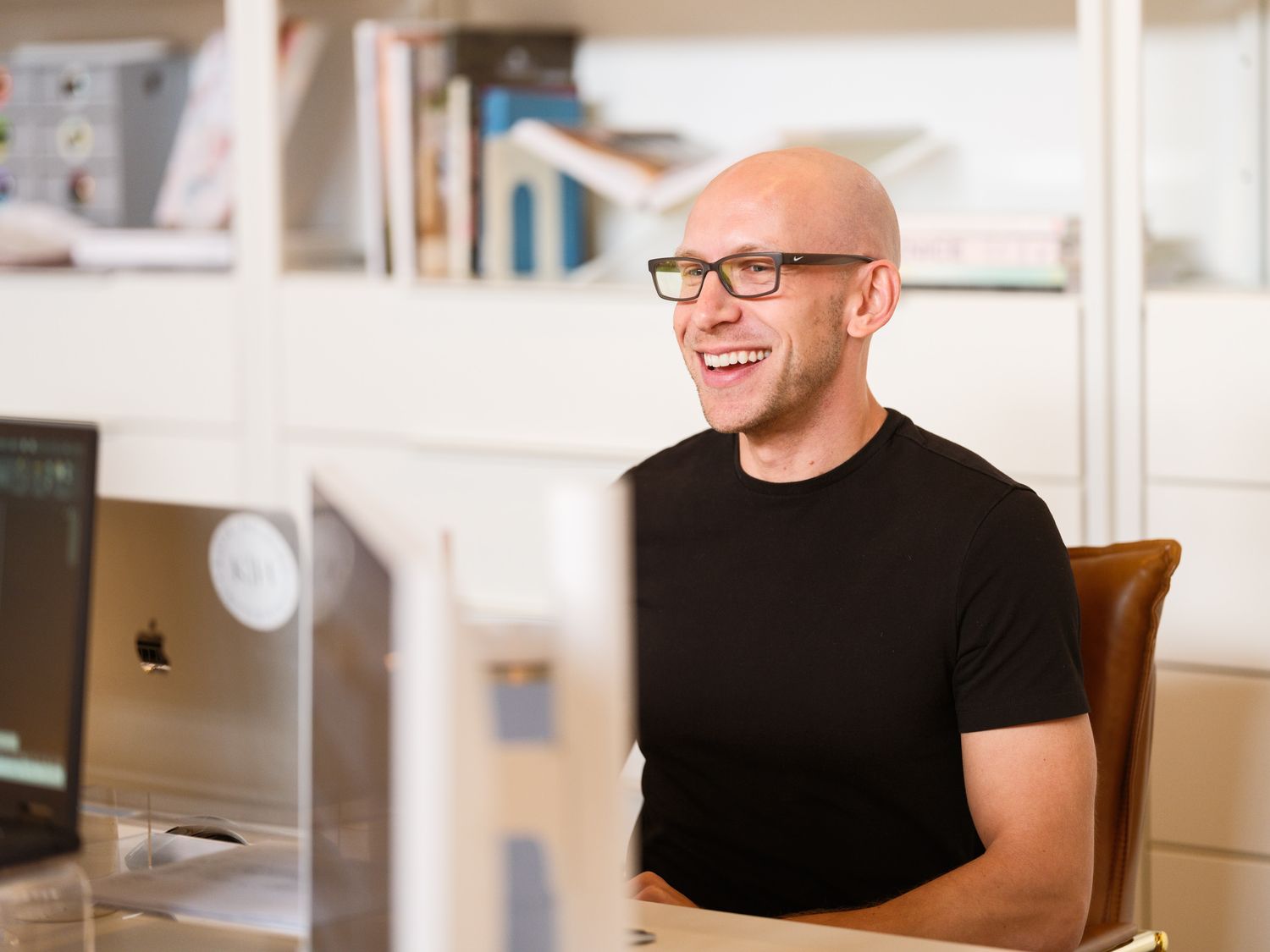 A smiling person in glasses and black t-shirt works at a desk with an Apple computer in a bright office space.