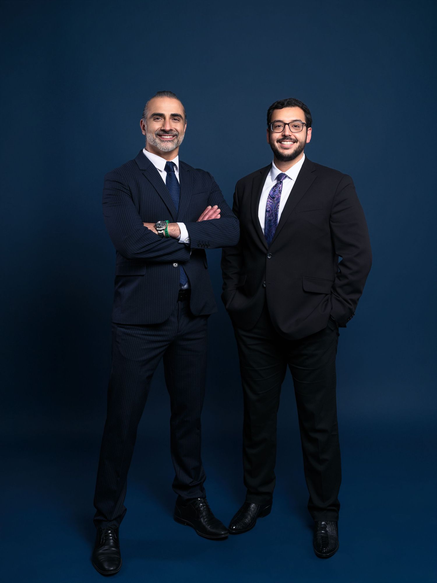 Two professionals in dark suits stand against a navy blue backdrop in a corporate photoshoot series.