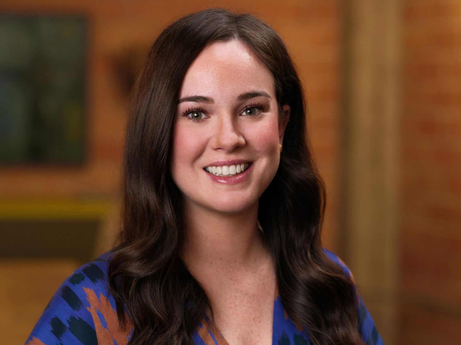 Business portrait of a woman wearing a blue patterned dress with long dark hair and a bright smile.