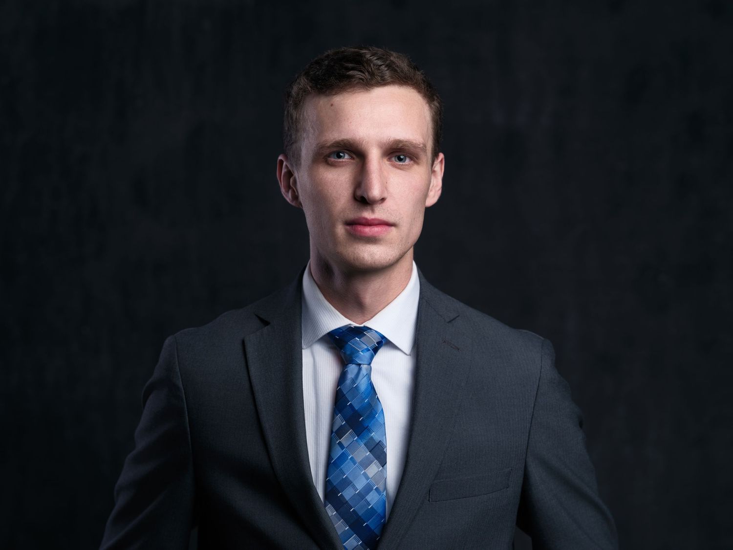 Young professional in dark suit with patterned blue tie against black background.