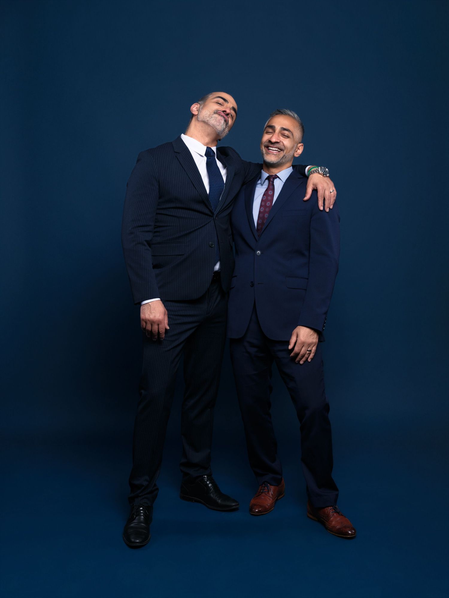 Two men in business suits share a lighthearted moment against a dark blue studio backdrop.