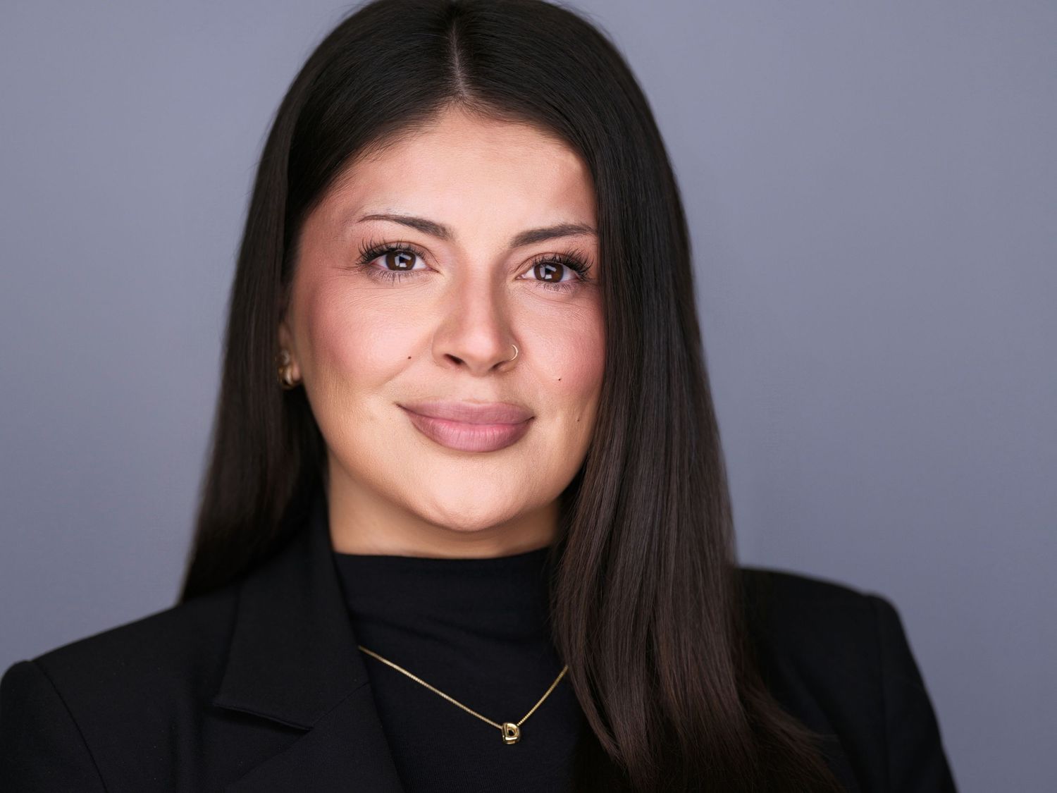 Professional headshot of a woman with long dark hair wearing a black blazer and gold necklace against a gray background.