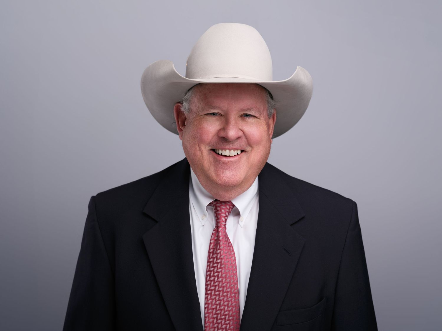 Professional in western cowboy hat wearing dark suit and red tie against gray backdrop.