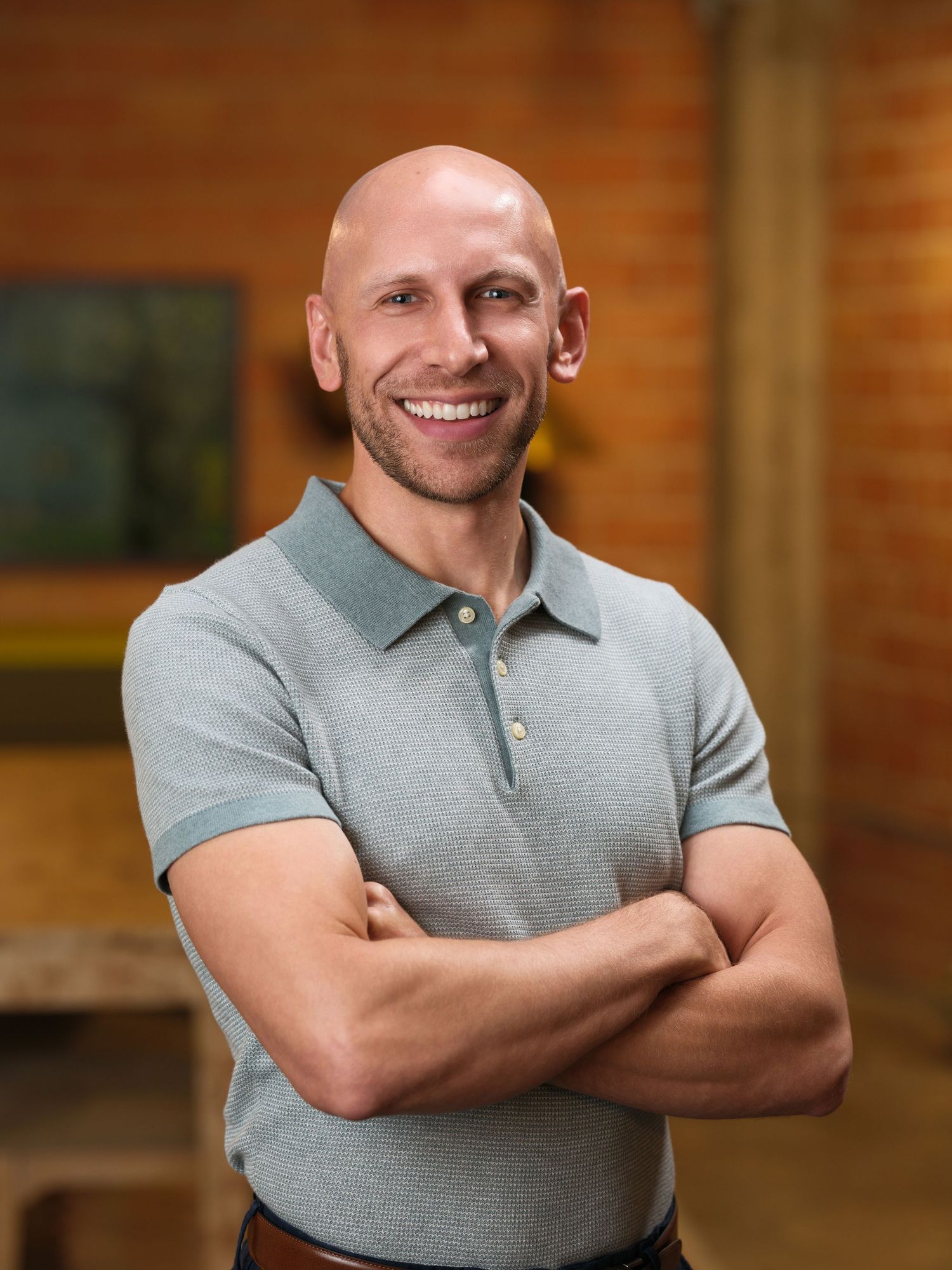 Person in gray polo shirt posing with crossed arms and warm expression against brick wall background.