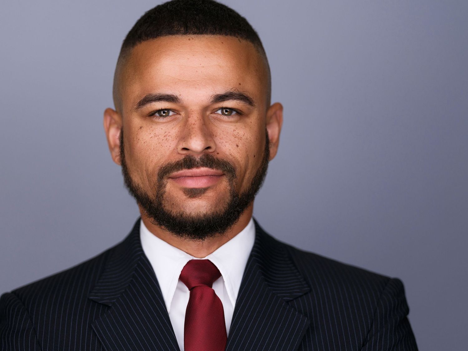 Professional headshot of a man in a dark pinstripe suit and red tie against a gray background.