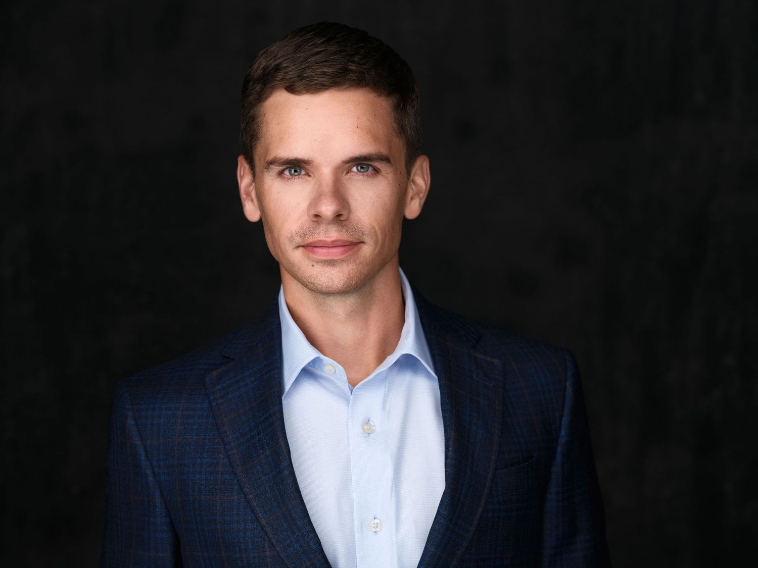 Professional headshot of a young businessman in a navy plaid blazer and light blue shirt against a dark background.