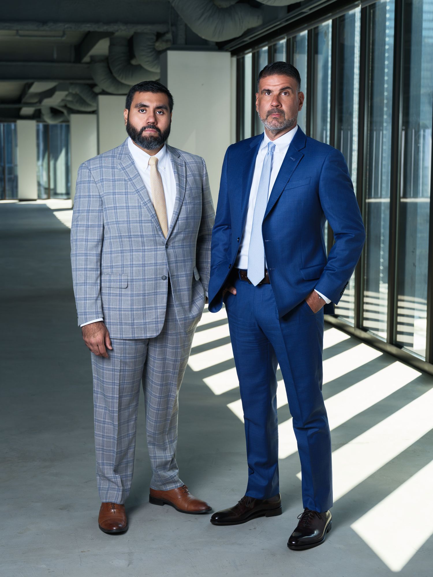 Two men in formal suits, one in gray plaid and one in navy blue, standing in a modern office hallway with sunlit windows.