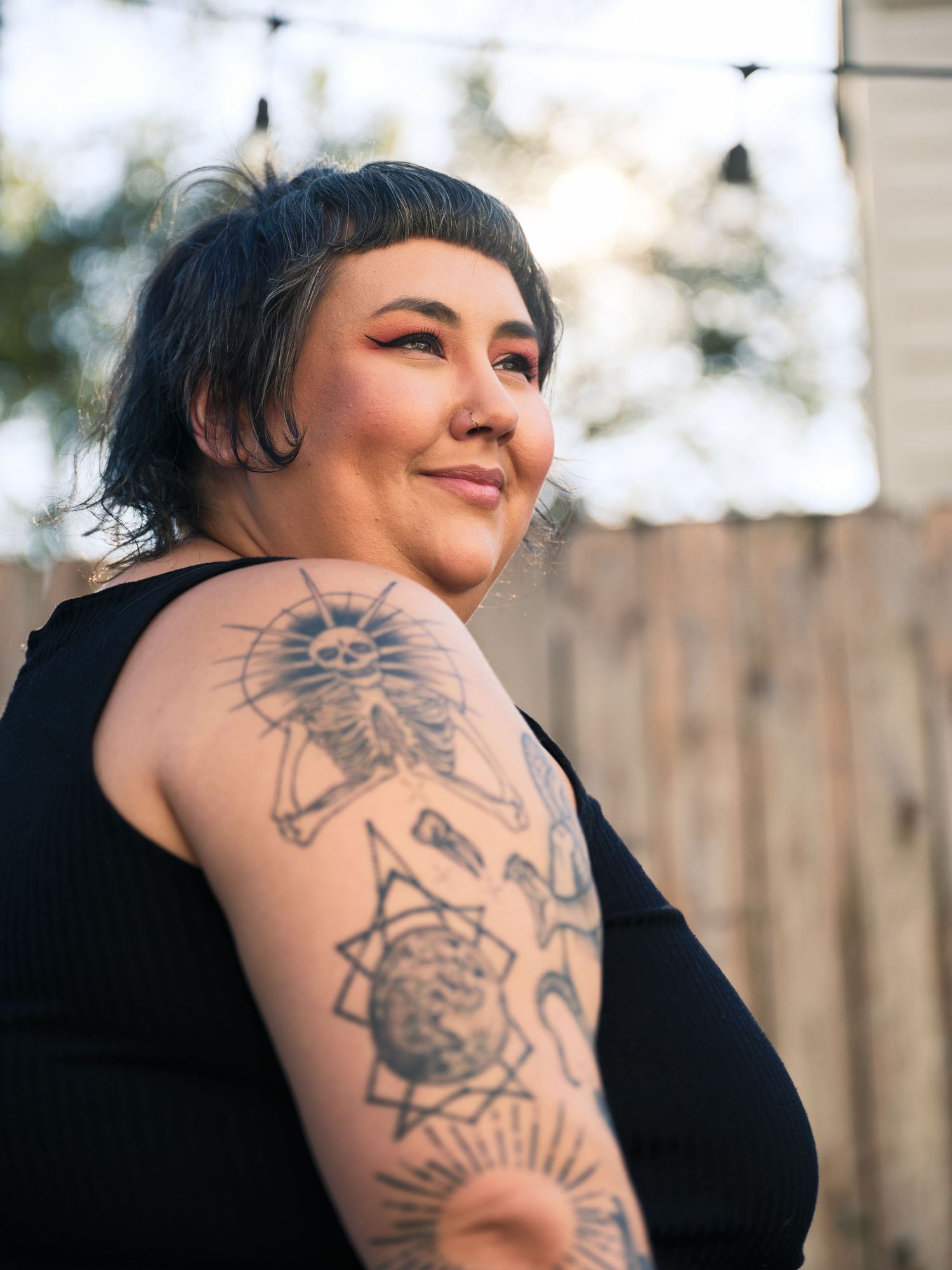 Person with dark hair and intricate arm tattoos poses outdoors in black sleeveless top.