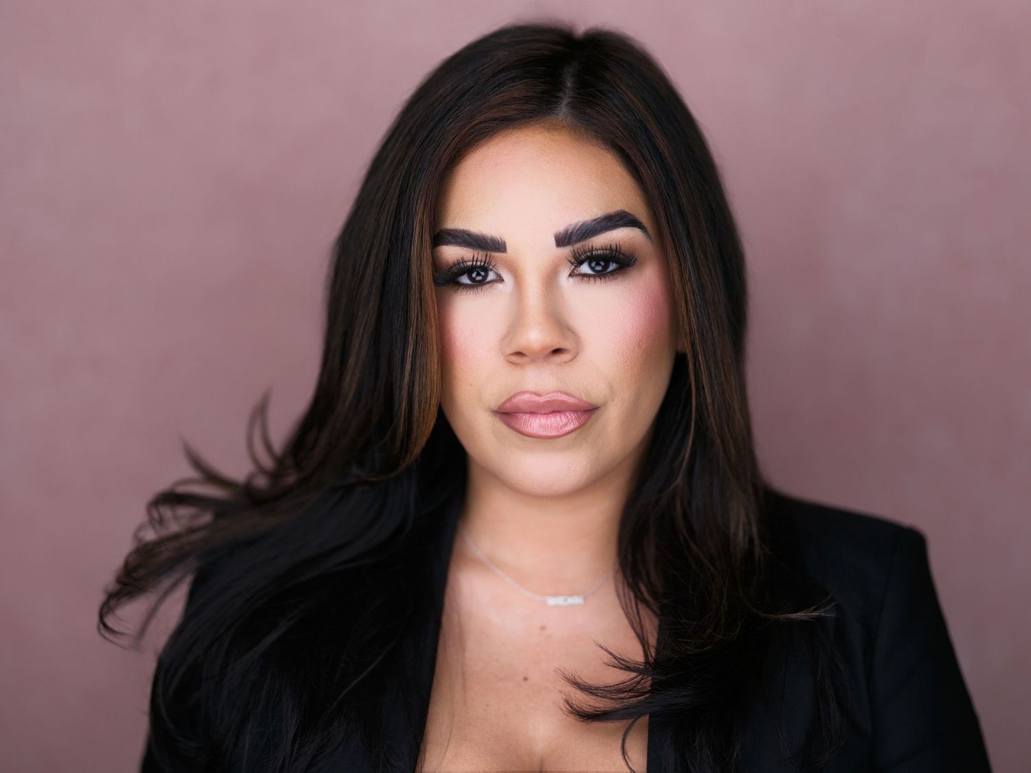 Professional portrait of someone wearing a black blazer against a pink background with glamorous makeup and dark hair.