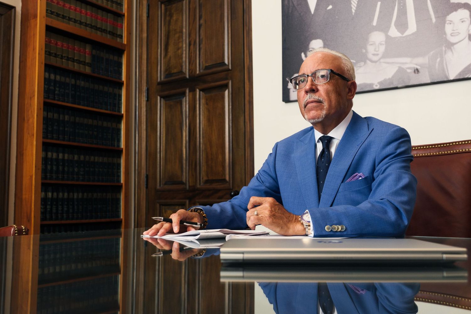 Senior legal professional in blue suit working at desk in law office with books behind him.