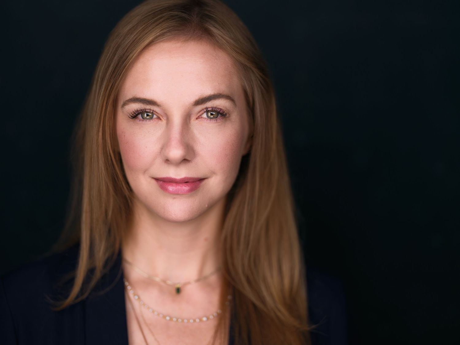 Professional headshot of a woman with long auburn hair wearing a navy blazer against a dark background.
