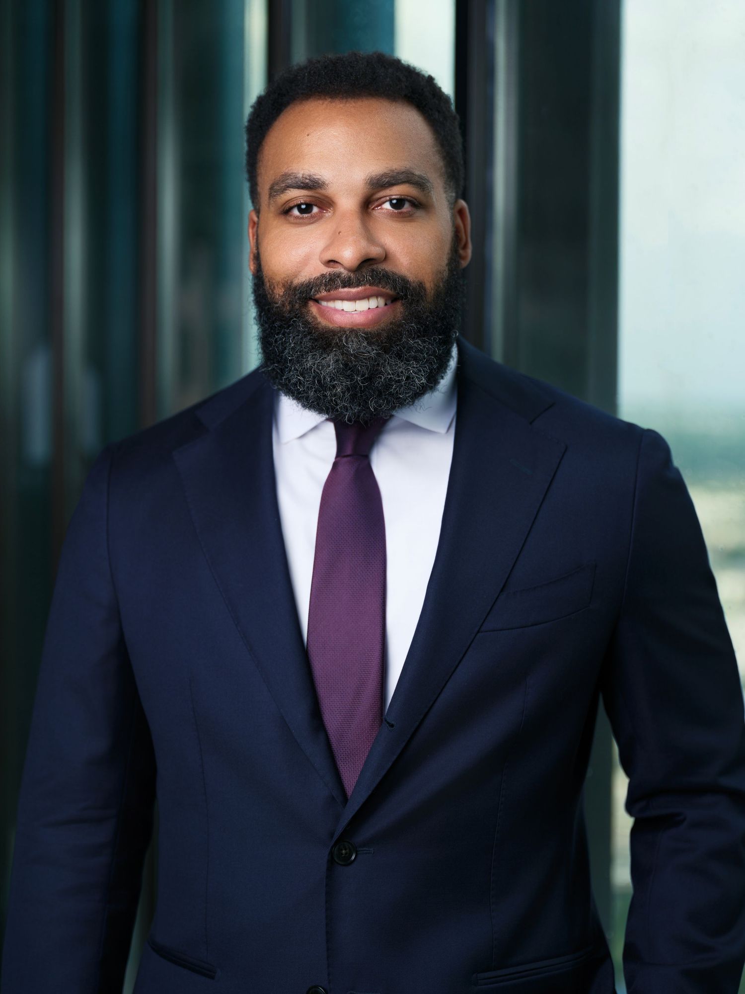Professional headshot of a businessman in a navy suit and purple tie with a full beard smiling at the camera.