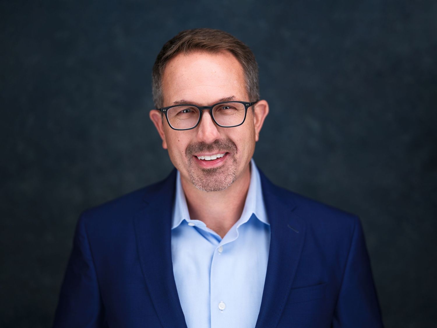 Professional man in navy blazer and glasses smiling confidently against dark studio background.