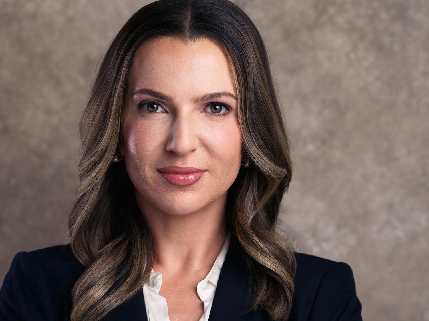 Professional headshot of a businesswoman with wavy brown hair wearing a navy blazer and white blouse against a neutral backdrop.