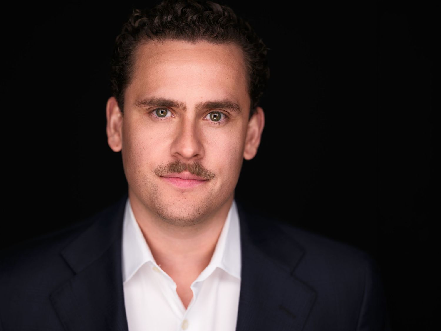 Professional headshot of a man with a mustache wearing a navy blazer and white shirt against a dark background.