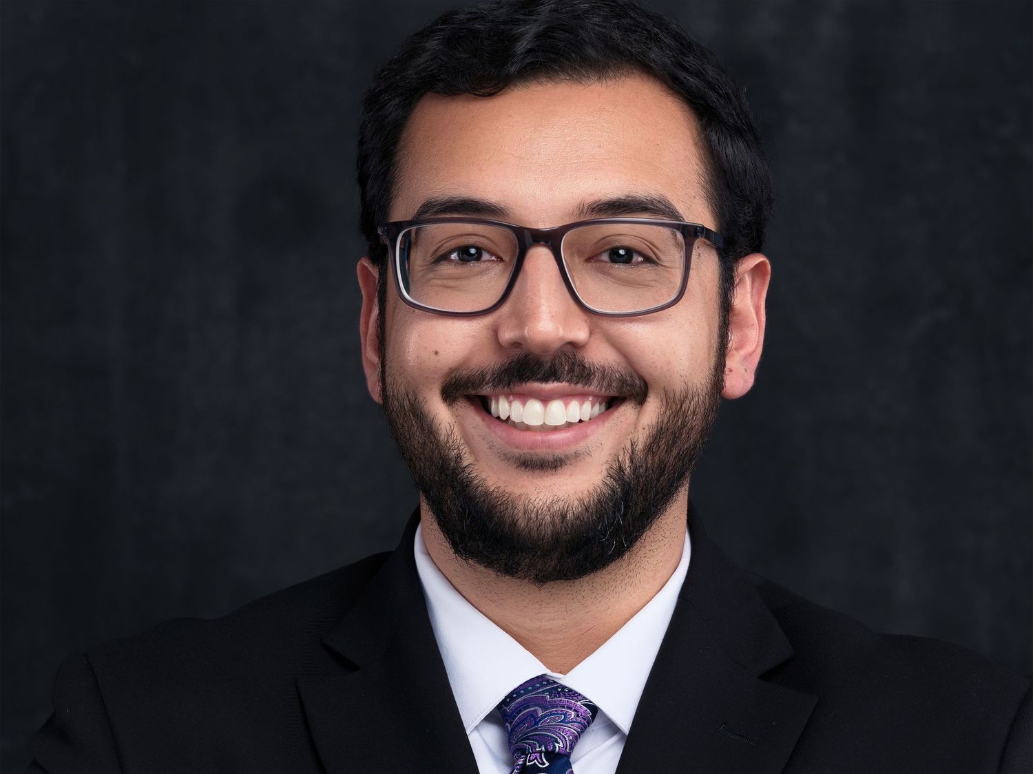 Professional headshot portrait of someone in a dark suit and glasses against black background showing a warm smile.