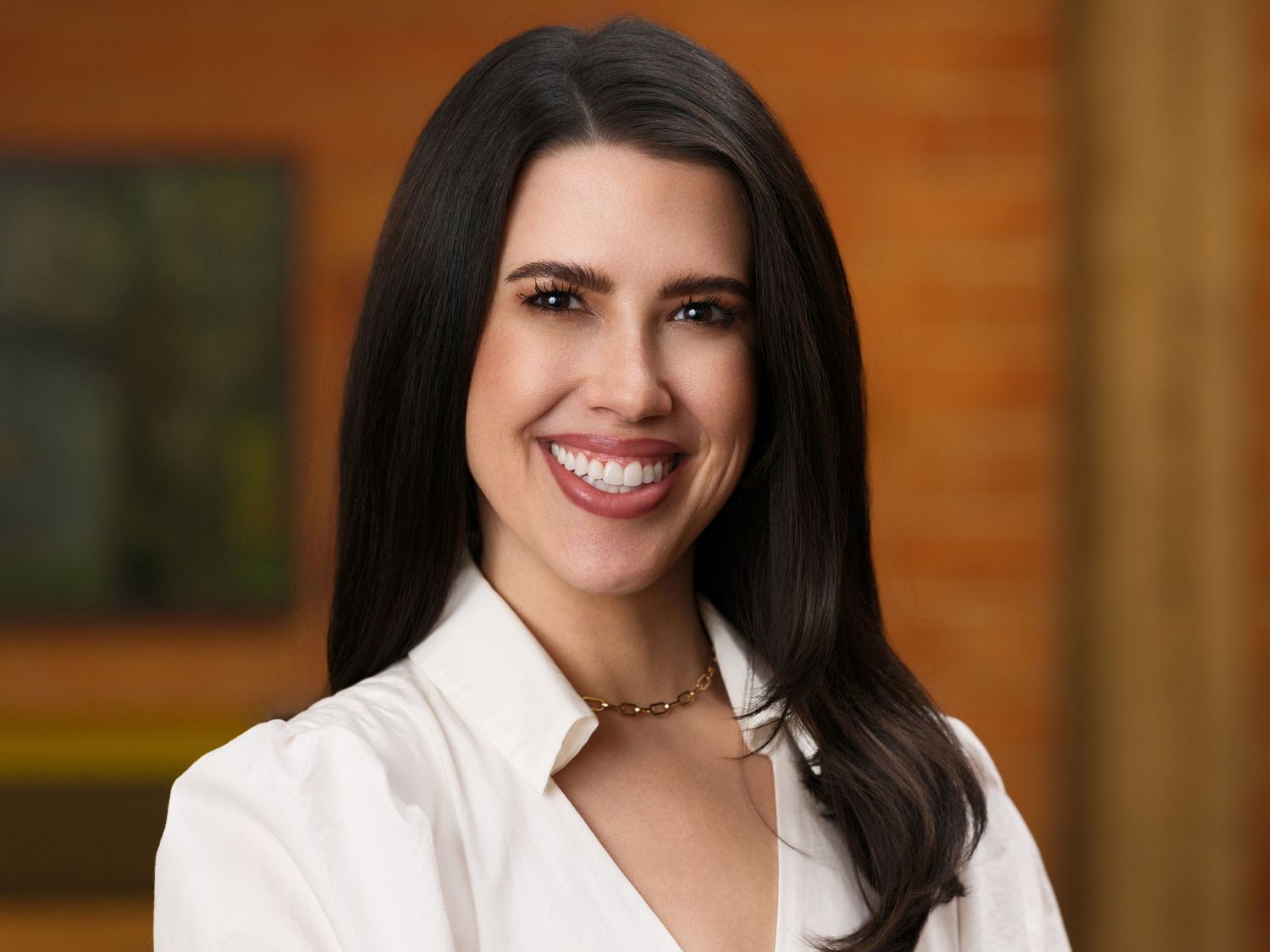 Professional headshot of a woman with long dark hair in a white blouse smiling warmly against a blurred background.