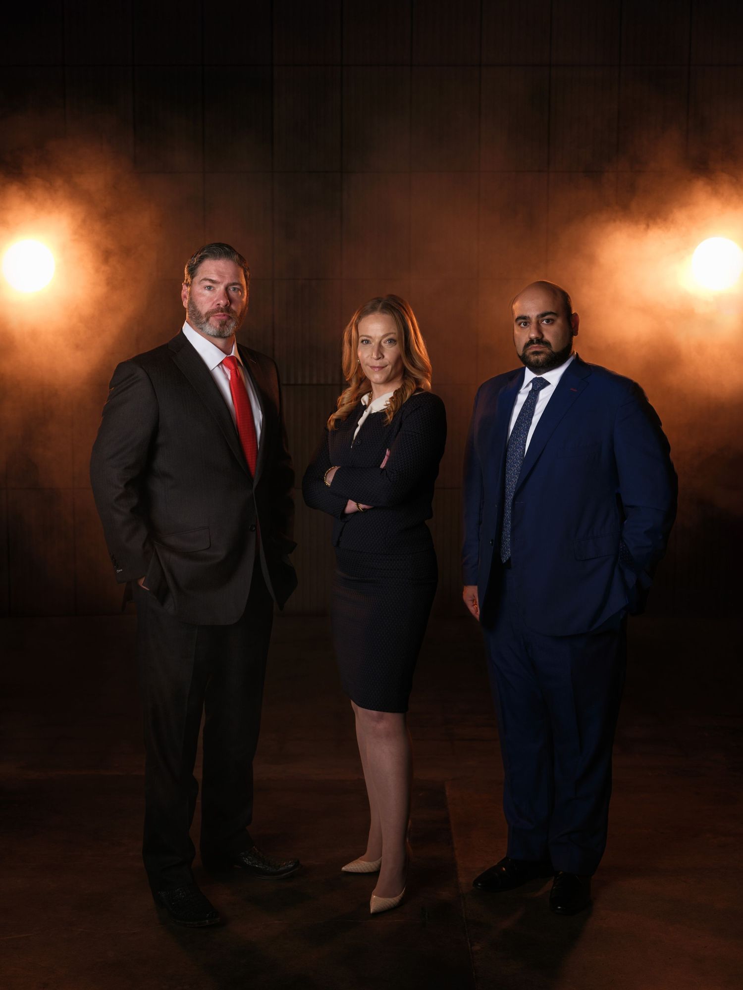 Three business professionals in formal attire posed dramatically with warm lighting in background.