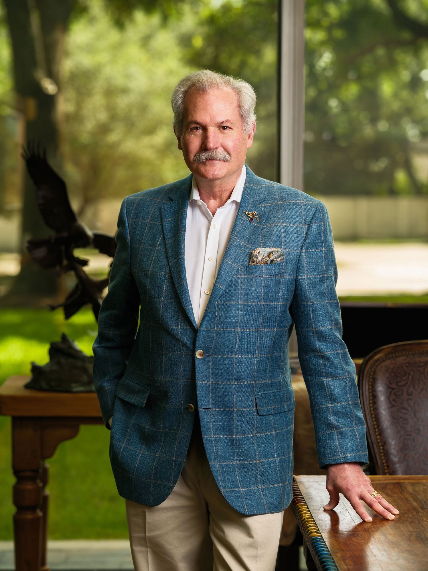 Distinguished older man with mustache wearing a blue plaid blazer poses confidently indoors near a wooden desk.