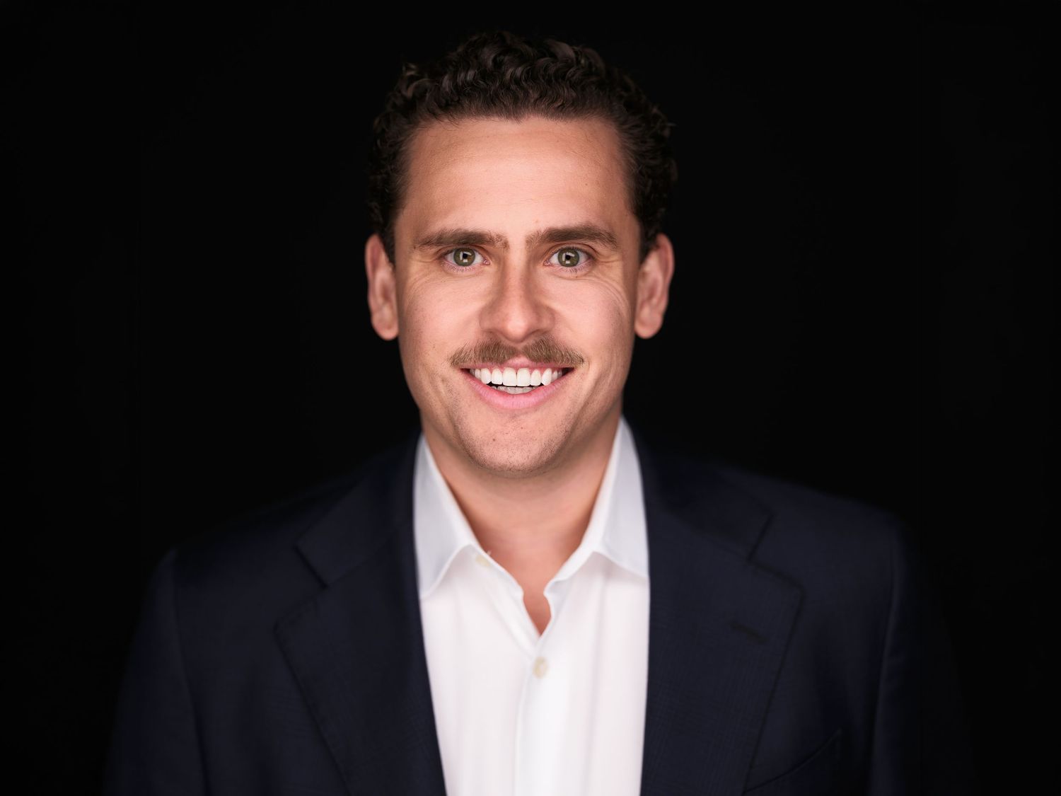 Professional headshot of a smiling man with a mustache wearing a navy blazer and white shirt against a dark background.