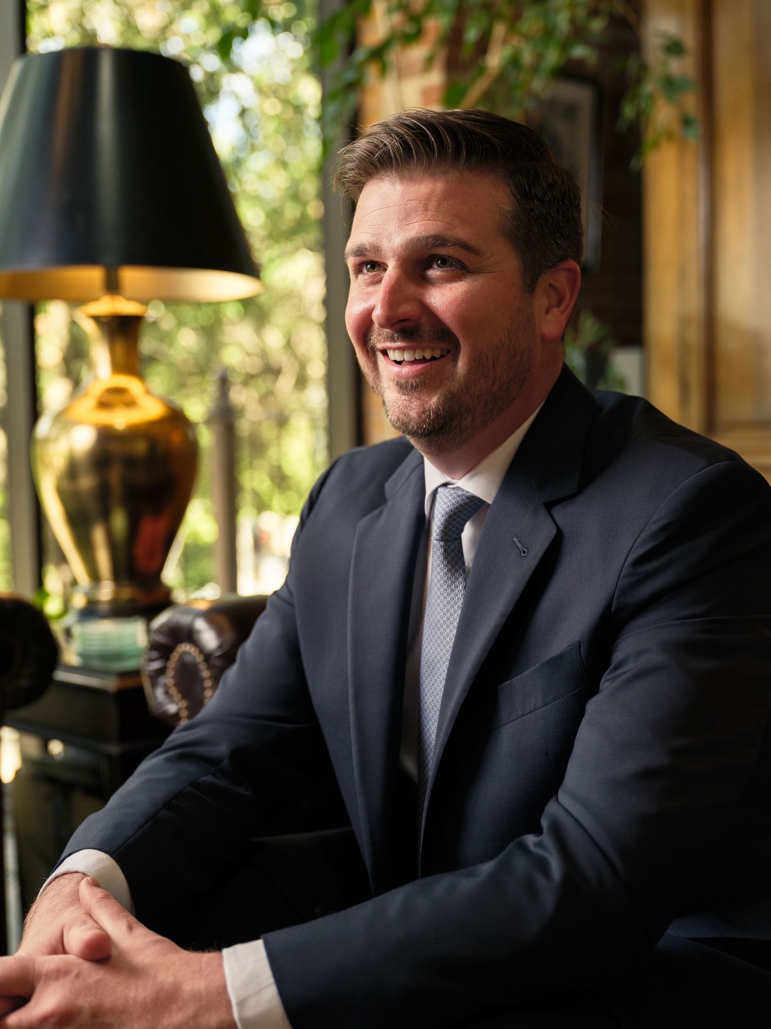 Professional portrait of smiling businessman in dark suit seated near decorative lamp and window.