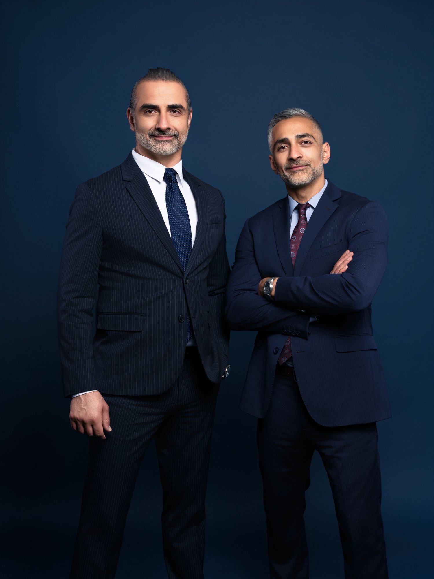 Two men in dark suits pose together against a navy blue background in a professional business portrait series.