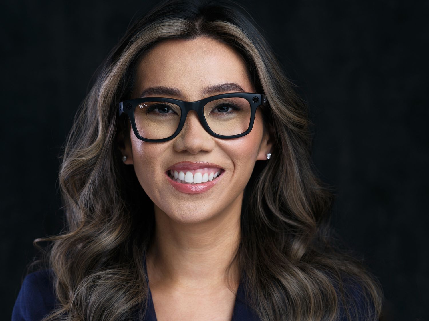 Professional headshot of a smiling person wearing black glasses and a navy blazer against a dark background.