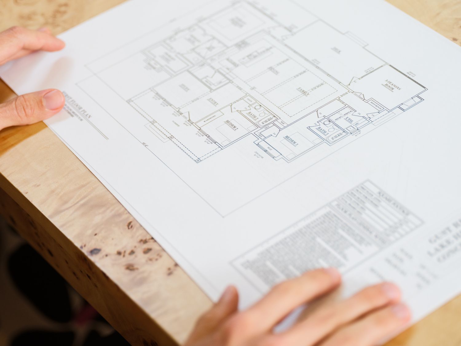 Hands holding architectural floor plan blueprints on wooden table showing detailed room layouts and measurements.