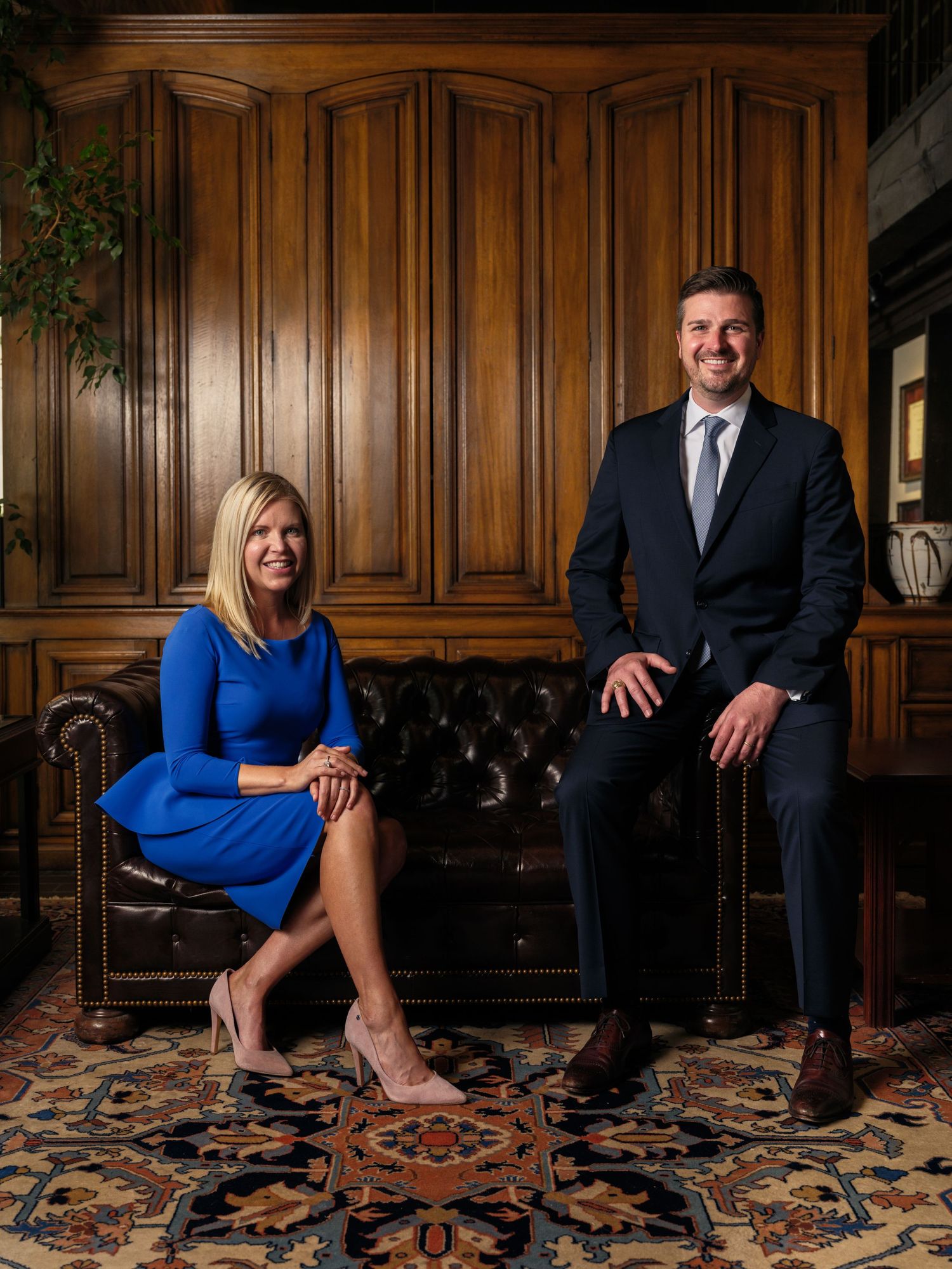 Corporate team photo with two professionals on leather sofa against wooden wall backdrop.