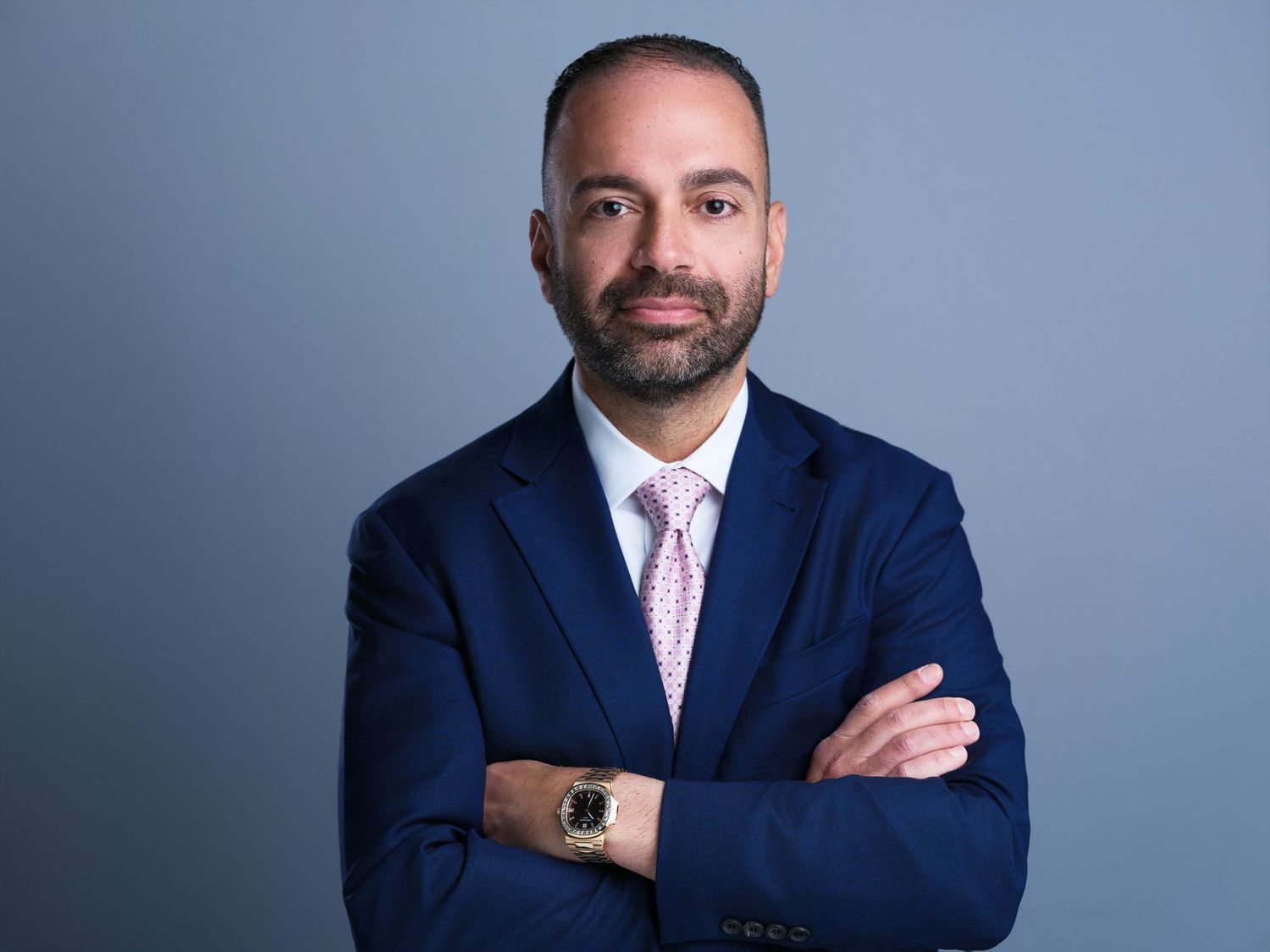 Professional headshot portrait of person in navy suit with arms crossed against blue studio background.