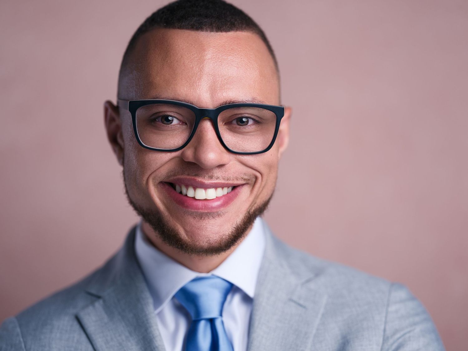 Professional headshot of someone wearing glasses, a blue tie and gray suit jacket against a pink background.