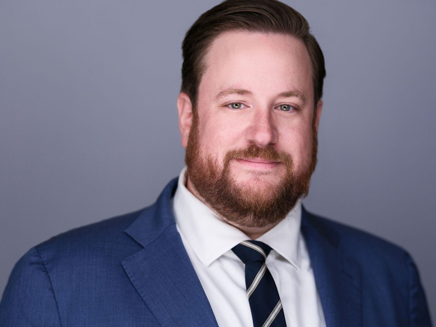 Professional headshot of a bearded man in a navy blue suit and striped tie against a gray background.