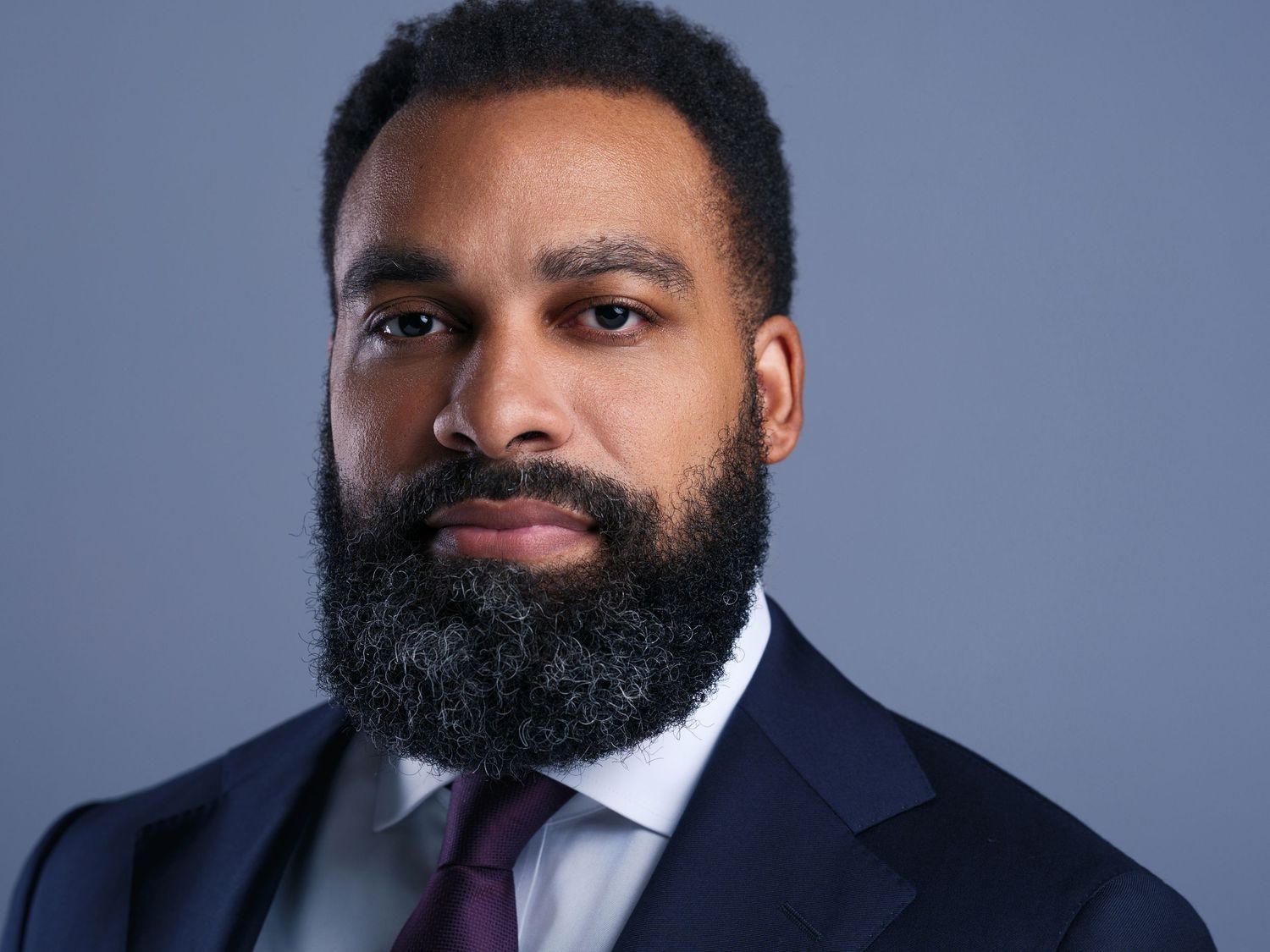 Professional headshot of someone wearing a navy suit and burgundy tie against a gray background.