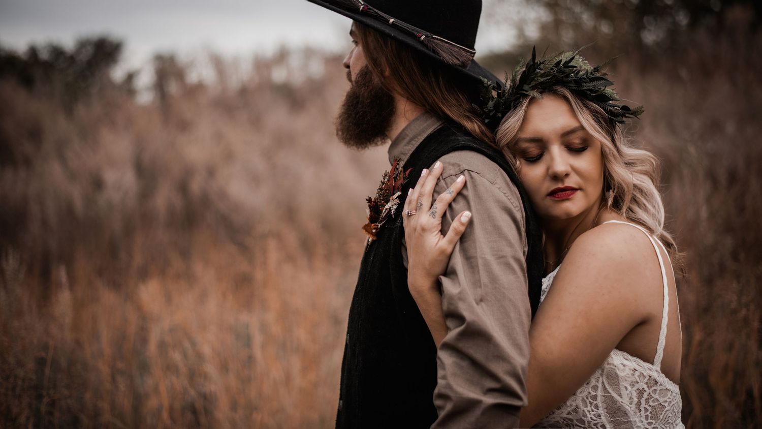 A romantic couple embraces in a golden field, with the person wearing a black hat and vest while their partner wears lace.
