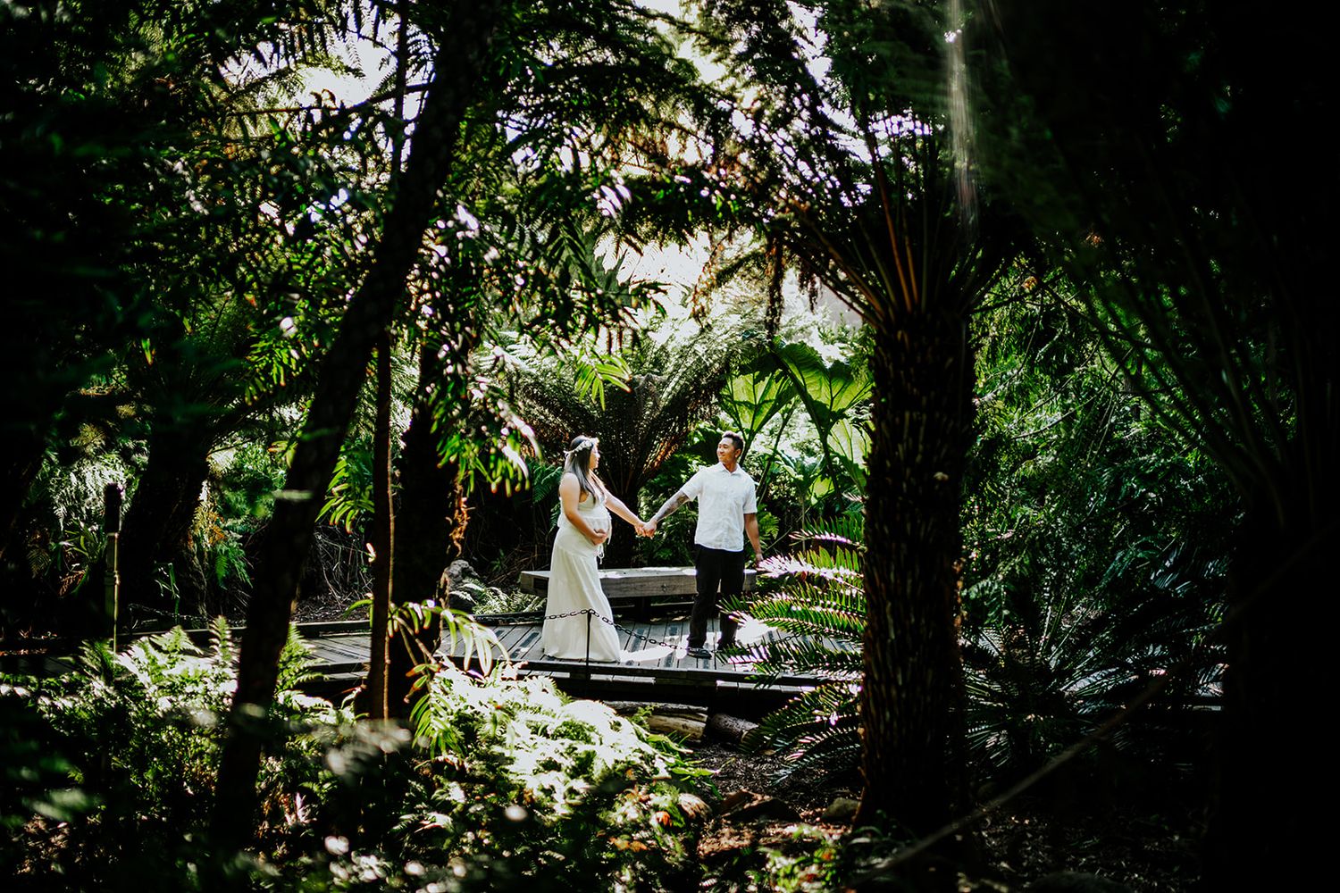 A couple holds hands on a bridge surrounded by lush tropical greenery in a romantic wedding photo.