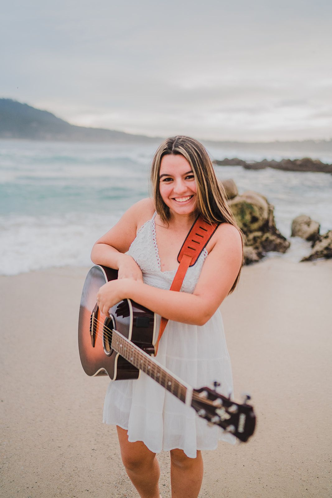 Smiling young woman in white dress holding acoustic guitar with brown strap on sandy beach with ocean waves.