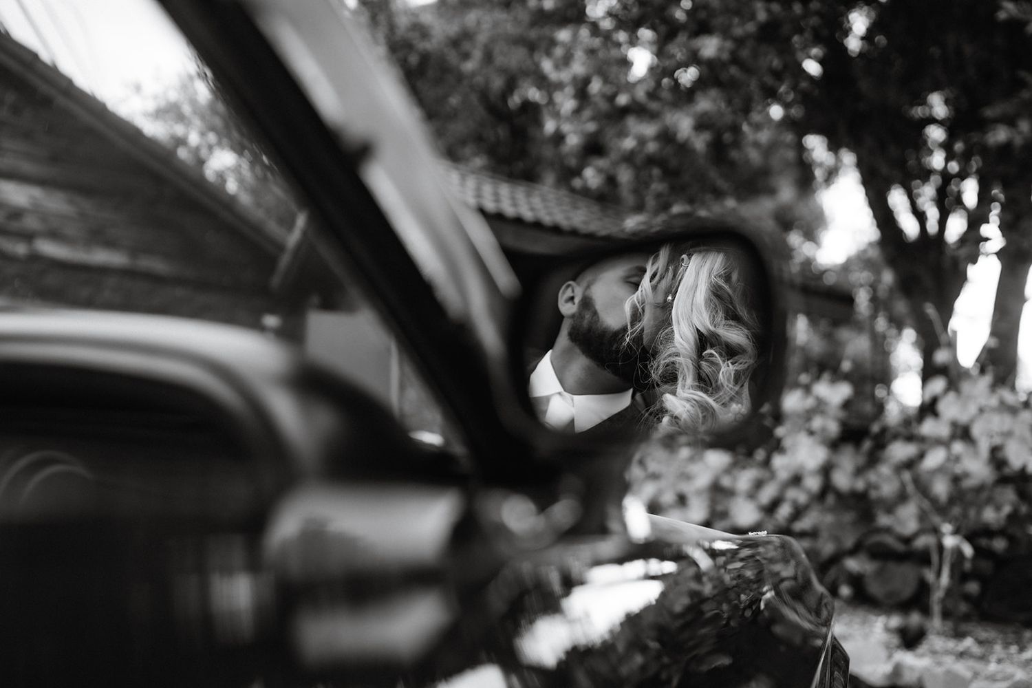 Black and white close-up shot through a car mirror reflecting trees and sky in soft focus.
