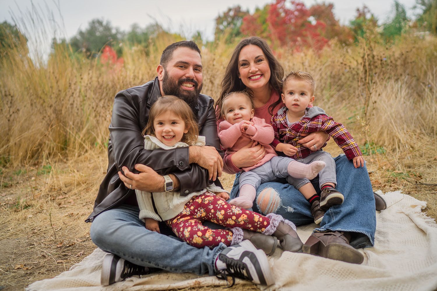 Two people sit casually on a blanket outdoors during fall with autumn colors and tall grasses in the background.