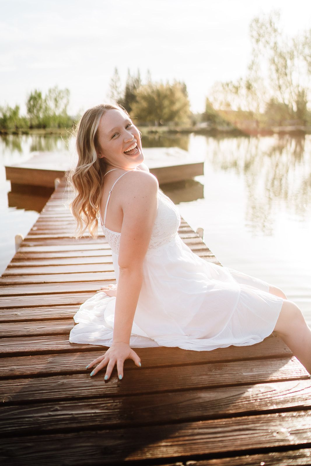 Smiling blonde woman in white sundress sitting on wooden dock by calm lake in golden sunlight.
