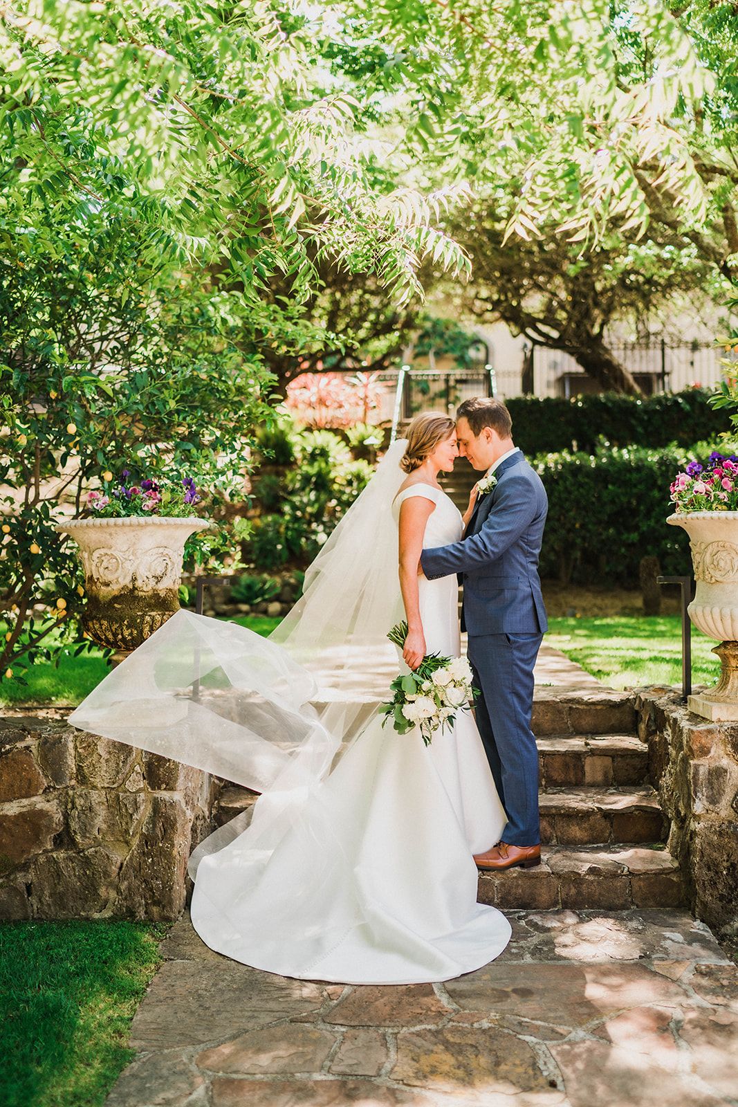 White wedding dress flowing in the breeze during outdoor garden portraits on stone steps.