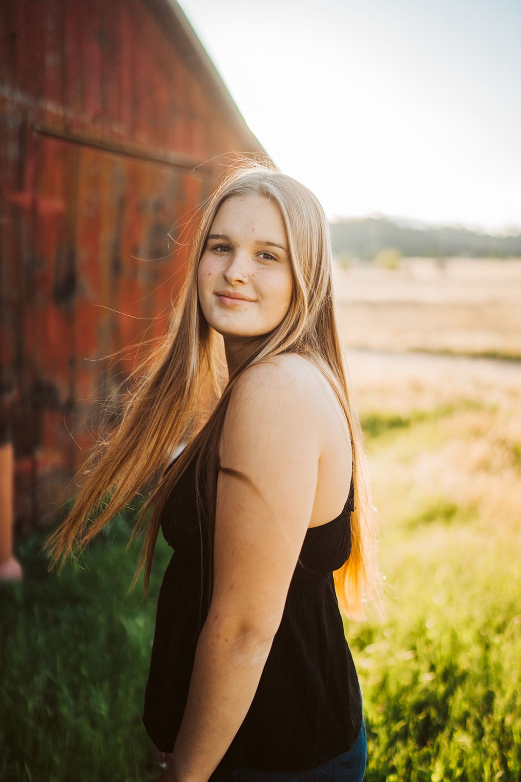 Young blonde woman in black tank top smiling outdoors near a red barn in golden sunset light.