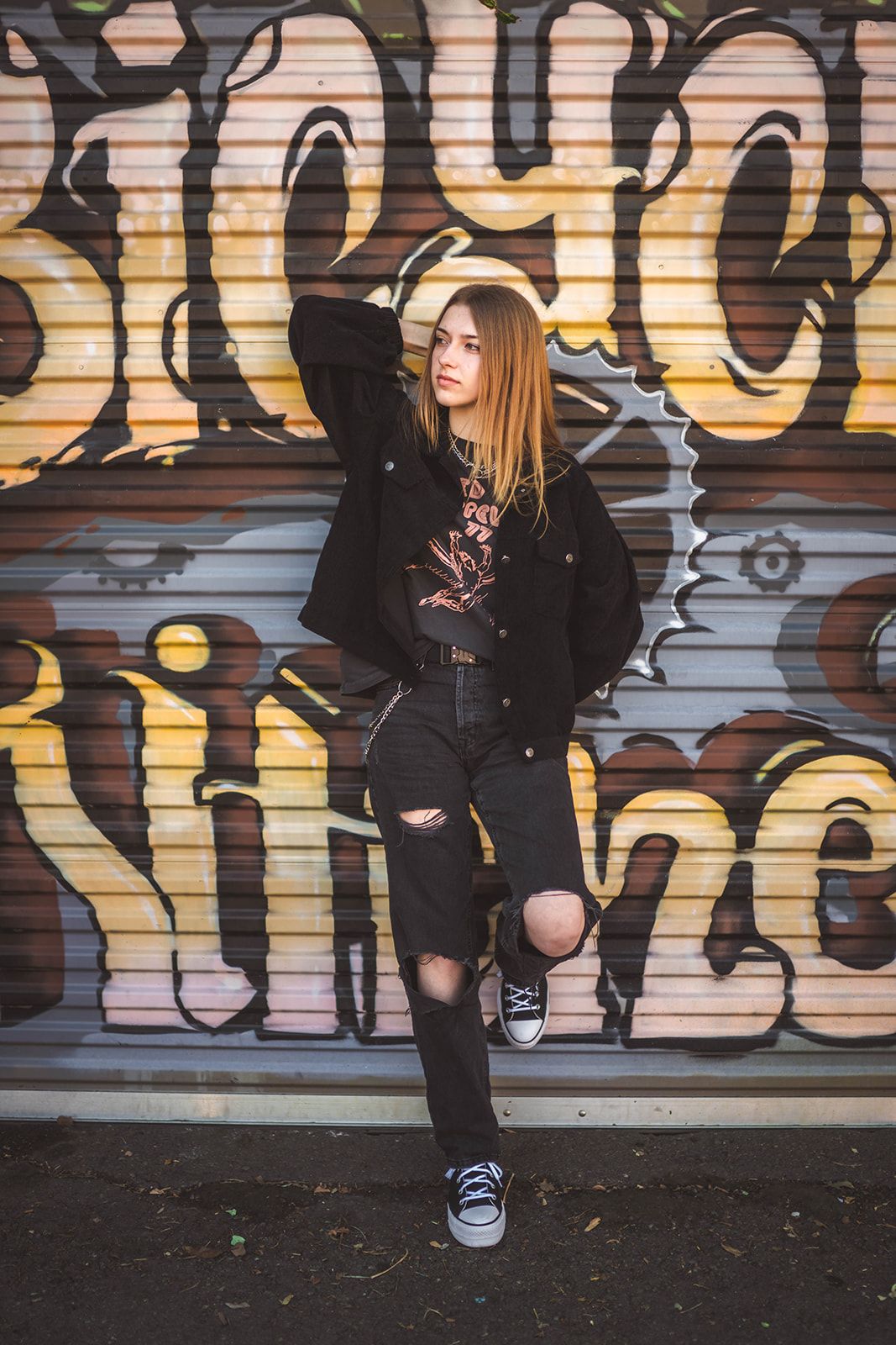 Young woman in black ripped jeans and jacket posing against colorful graffiti wall in urban street style.