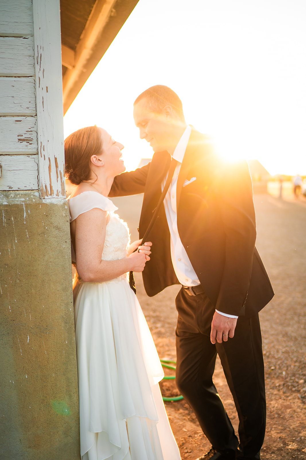 Bride and groom share a romantic golden sunset moment leaning against a rustic white pillar in wedding attire.