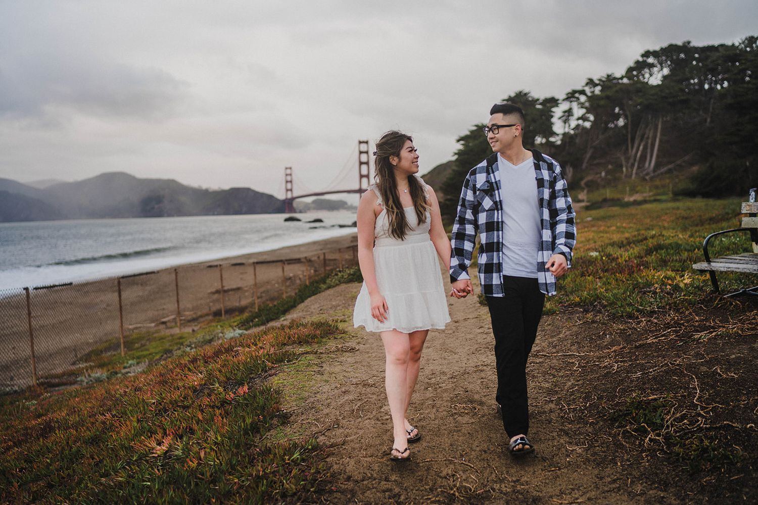 Couple walking along coastal path with Golden Gate Bridge visible in background.