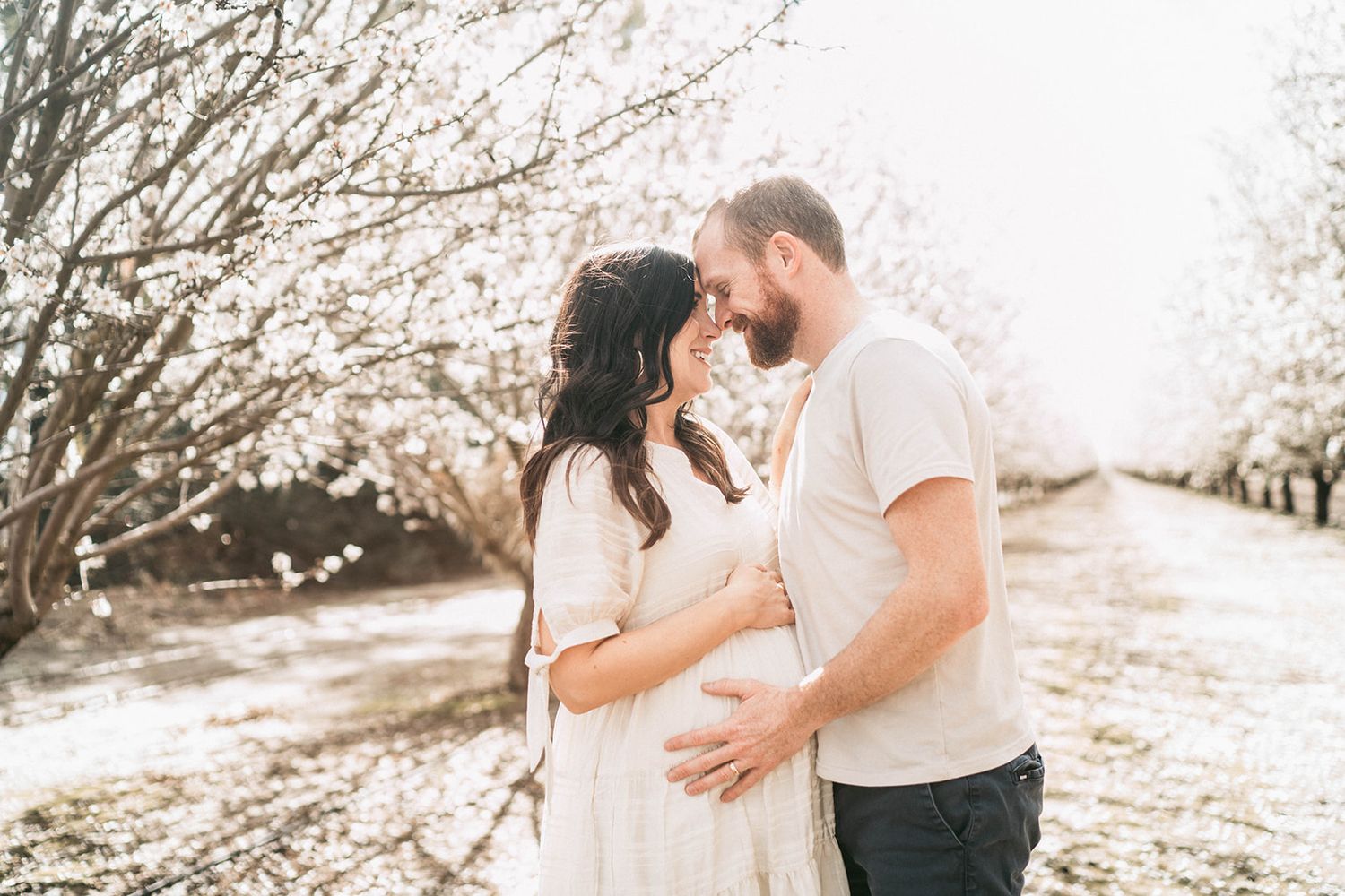 Couple embracing tenderly in blooming orchard, maternity photo shoot among white spring blossoms.