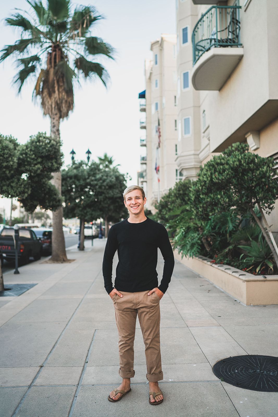 Smiling young man in black sweater and khaki pants stands on a palm-lined urban sidewalk.