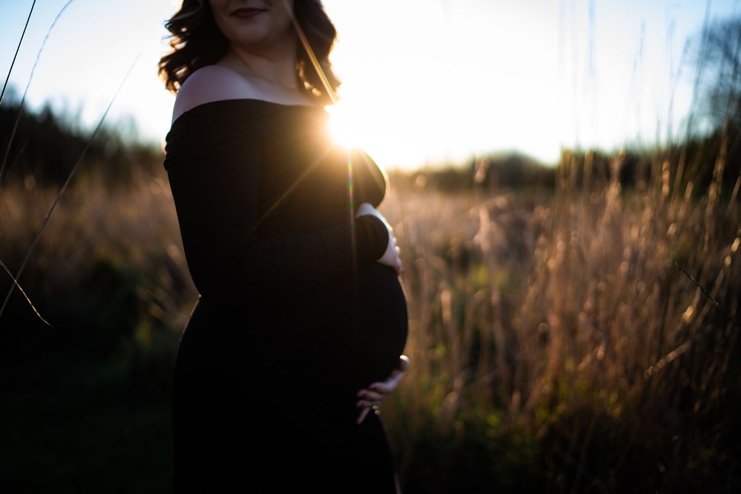 Pregnant woman in black off-shoulder dress cradling baby bump at golden sunset in a field of tall grass.