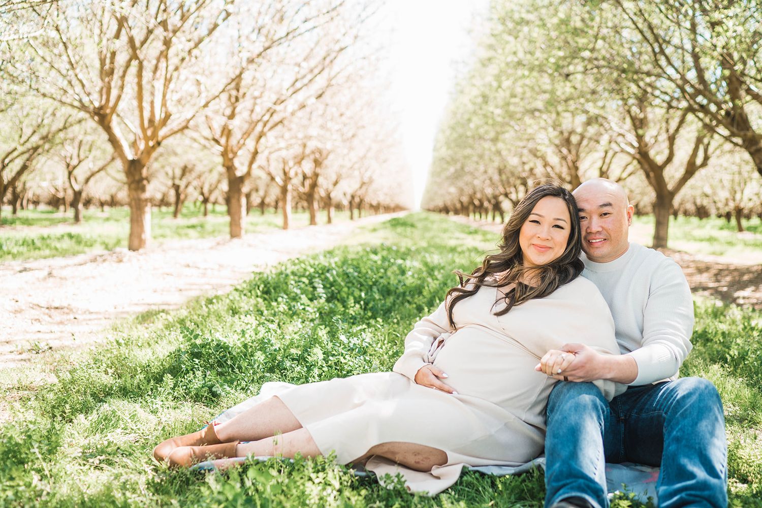 Couple sitting in blooming orchard, pregnant woman in white dress, man in grey sweater, spring maternity photo.